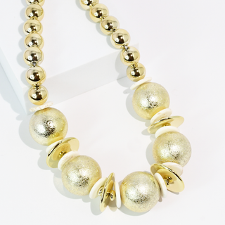 A gold beaded statement necklace with textured metallic gold beads, matte gold disc beads, and small cream-colored spacer discs.