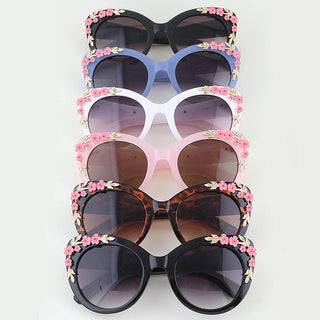 Five pairs of oversized cat-eye sunglasses. The frames are in black, blue, white, pink, and tortoiseshell, each decorated with pink flowers and gold leaves around the rims.