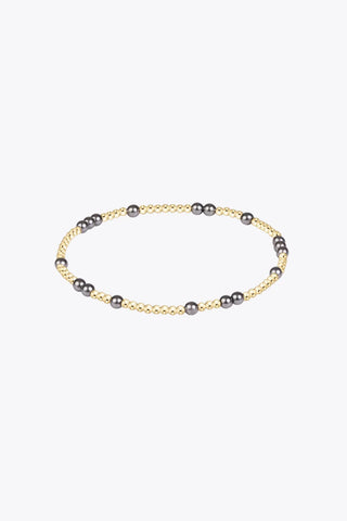 A stretch beaded bracelet featuring small polished 14K gold-filled beads interspersed with larger dark grey pearl crystal beads by Enewton.