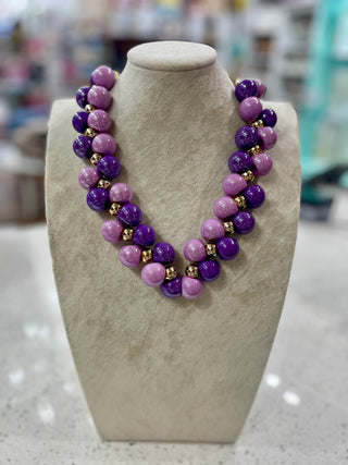 A chunky beaded necklace displayed on a beige fabric jewelry bust. The necklace features alternating strands of polished purple and lilac beads, separated by small metallic gold spacer beads.