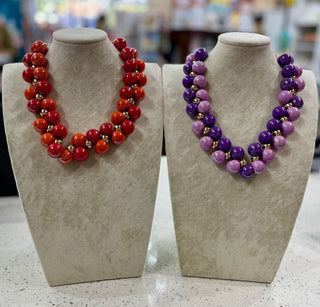 Two beaded statement necklaces displayed on beige jewelry busts. The necklace on the left features round red and orange beads with small metallic spacer beads and the necklace on the right has purple and lilac beads.