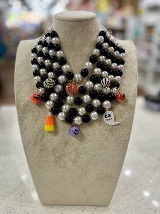 A multi-strand beaded necklace displayed on a beige fabric mannequin bust. The necklace features strands of black and white beads with small colorful charms, including a candy corn, a ghost, and a purple pumpkin.