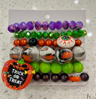 A set of Halloween-themed beaded bracelets featuring purple, green, orange, marbled black-and-white beads, a gummy eyeball, lime green beads, dark purple beads, and a decorative orange pumpkin sign that reads 'Trick or Treat'.