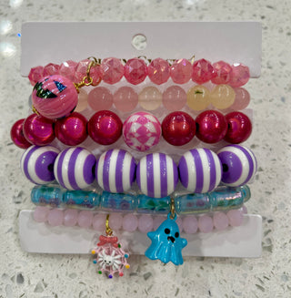A set of six colorful beaded bracelets. The bracelets feature pink faceted beads, magenta glossy beads, purple and white striped beads, translucent blue beads, and a light pink beaded bracelet with a blue ghost charm and a pink snowflake charm.