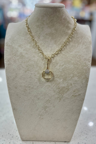 A gold chain necklace with a snaffle bit pendant featuring a gemstone, displayed on a beige velvet jewelry bust.