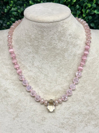A pink beaded necklace displayed on a cream-colored jewelry bust. The necklace features faceted light pink beads, smaller round pink beads, and a central clear teardrop-shaped crystal pendant with a gold-toned setting.