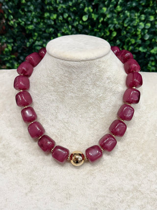 A chunky beaded necklace made of glossy, rounded magenta-pink beads separated by small gold spacer rings, featuring a larger central gold bead with a floral motif. The necklace is displayed on a textured cream-colored jewelry bust.