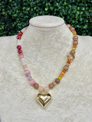 A beaded necklace with a polished gold heart pendant, displayed on a textured cream jewelry bust. The necklace features multiple strands of beads in pink, peach, brown, and red, accented with small gold spacers.