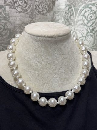 A classic white pearl necklace displayed on a cream-colored fabric jewelry bust, worn over a black top.