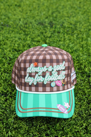 A baseball cap with a brown and beige gingham patterned crown and a mint green striped visor, placed on a green grass background. The front of the cap features the text 'always a good day for football' in white script with a green outline, decorated with small football-themed patches.