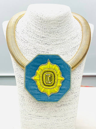 A chunky gold-tone collar necklace with a large blue octagonal pendant featuring a yellow sunburst design and a central green gemstone.