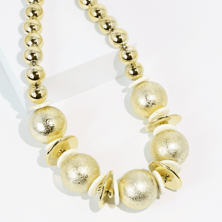 A gold beaded statement necklace with textured metallic gold beads, matte gold disc beads, and small cream-colored spacer discs.