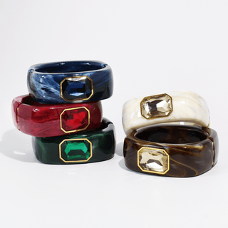 Five chunky resin bangles with large faceted gemstone accents, stacked on a white background. The bangles feature marbled patterns in navy blue, red, emerald green, and dark brown, with cream and clear stones accented by gold-tone bezel settings.