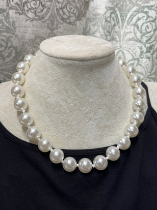 A classic white pearl necklace displayed on a cream-colored fabric jewelry bust, worn over a black top.